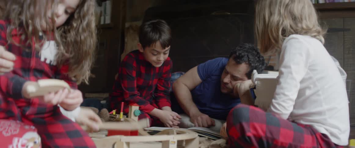 A father is reading a book with a boy as a family play on a living room floor