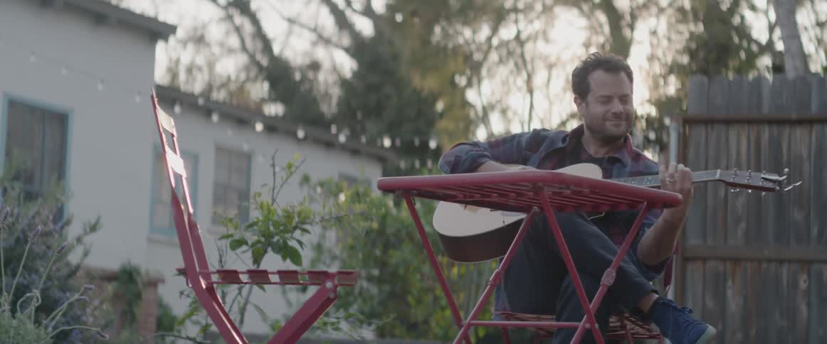A man is playing guitar and watching a boy ride a bike in a backyard in daylight
