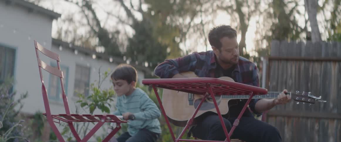 Man plays the guitar outside while a child rides a toy bike