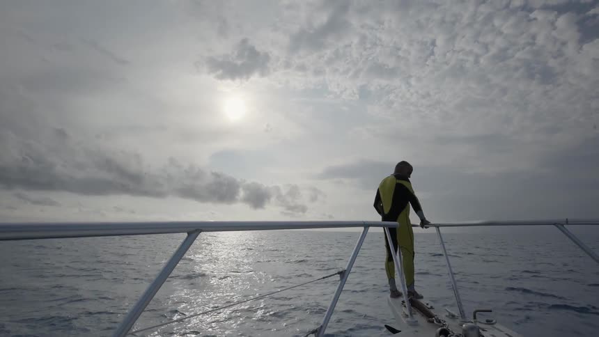 Man standing on the very front railing of a boat moving towards a cloudy sunset