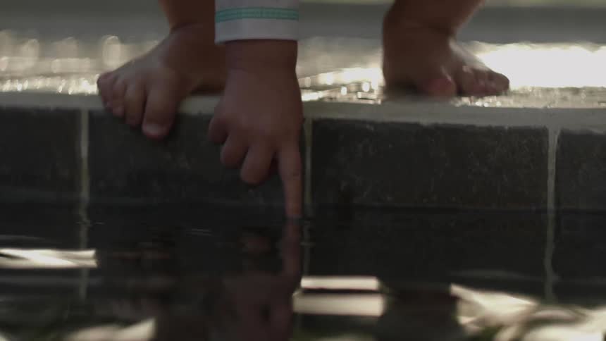A child is reaching off the edge of a pier to touch some water outside in daylight
