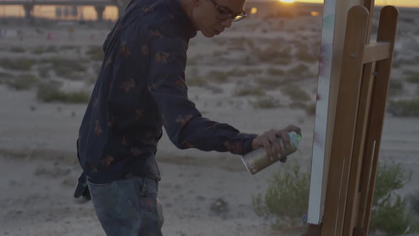 Man spray paints on a canvas while standing in a field
