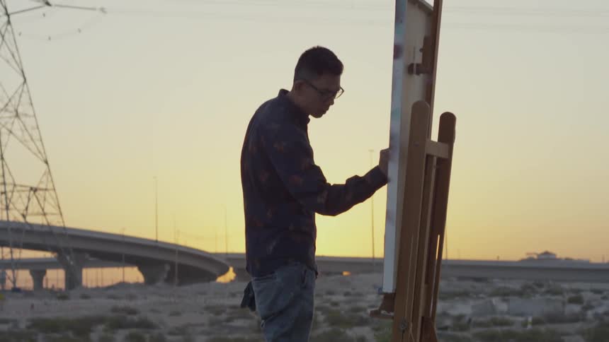 An artist paints on a canvas in a desert at sunset