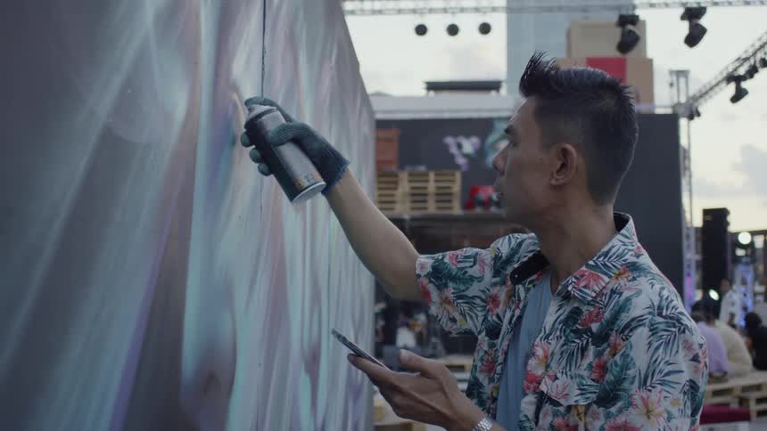 A man looking at his phone while spray painting a wall