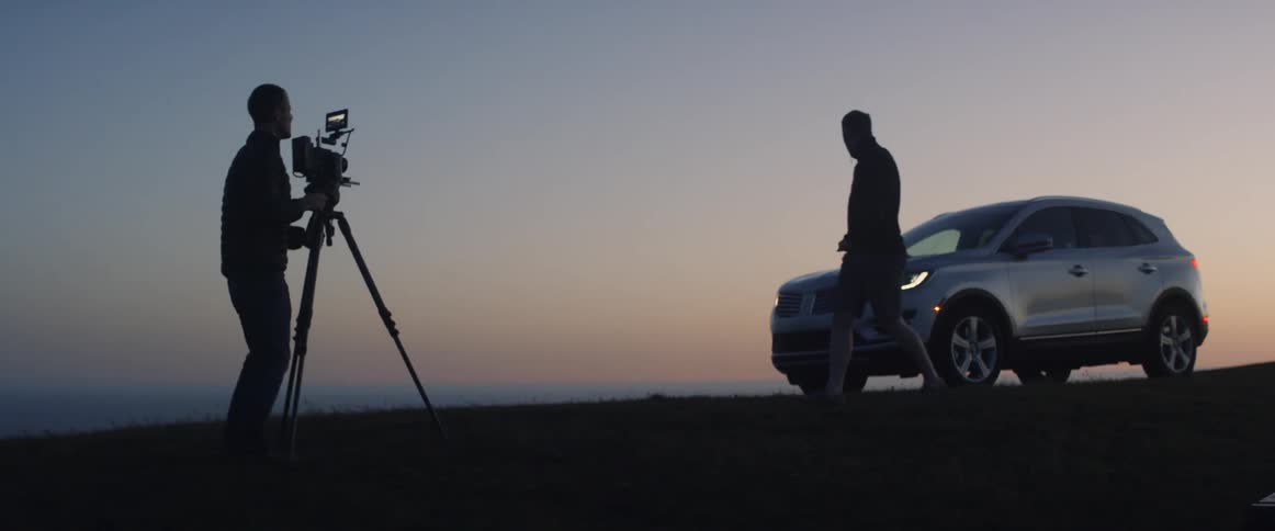 Person carrying a tripod walks towards a person and a car at sunset