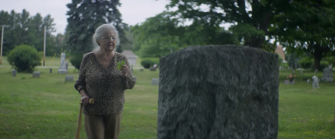 An elderly woman stands in front of a headstone in a cemetery