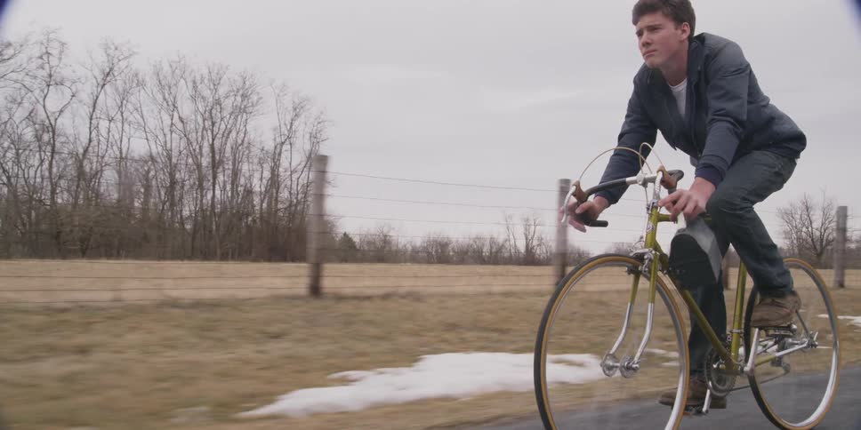 A man holding a lunch box and riding a ten speed down a rural area that still has patches of snow alongside the road