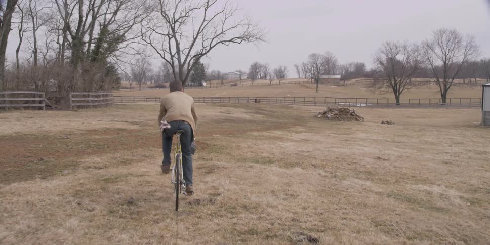 Man riding his bike across a field holding a box