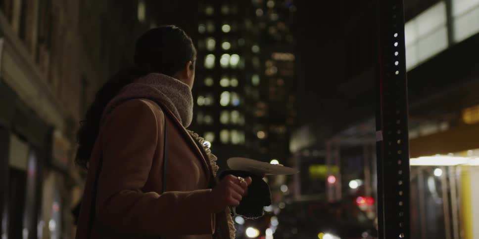 Woman on urban sidewalk eats pizza slice at night