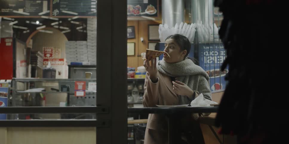 A woman is eating a slice of pizza inside a fast food restaurant at night