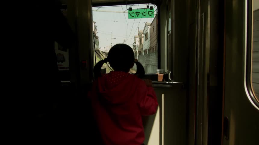 A girl is watching out a train window as it travels through a neighborhood in daylight