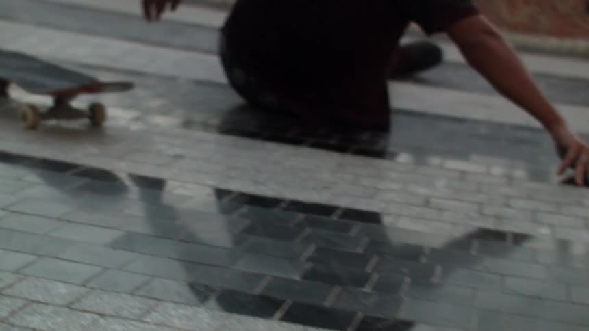 A man riding a skateboard near a fountain in daylight stumbles and falls