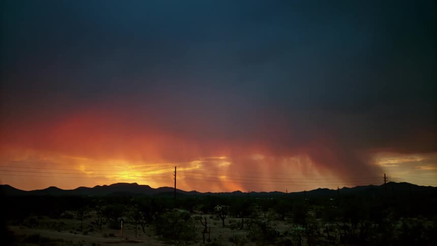 Lightning is flashing amid storm clouds as they move across a landscape at sunset