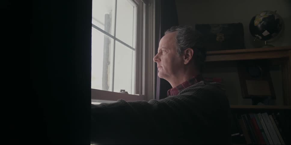 An older white man stands at a window and looks outside