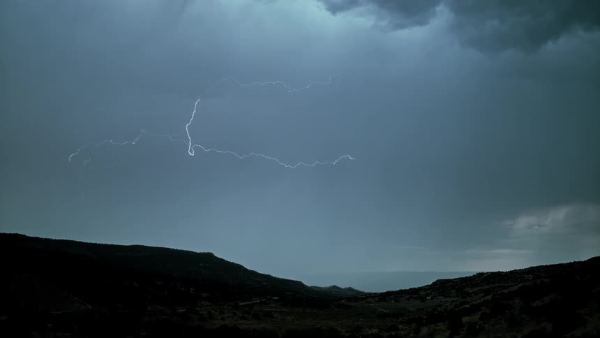 Lightning is flashing amid storm clouds as they move across a landscape at dusk