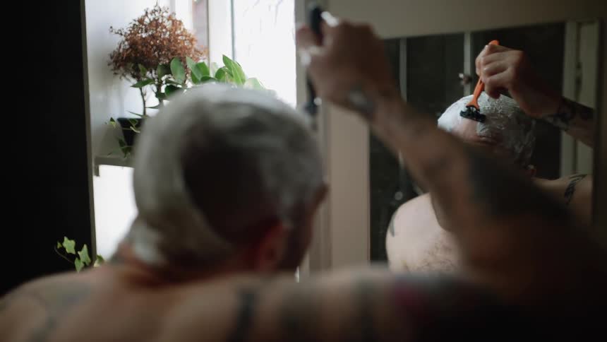 A man is shaving his head as he stand in a bathroom in daylight
