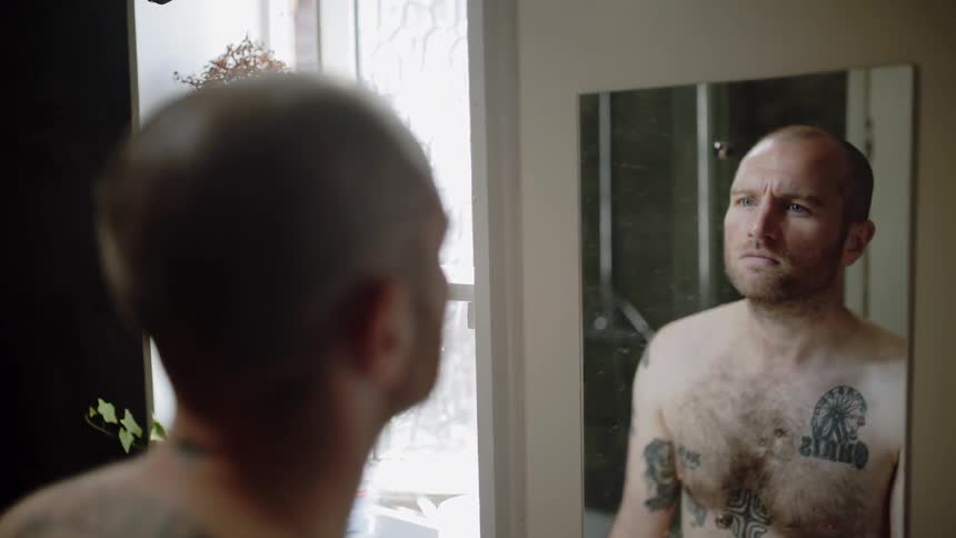 A shirtless man looks in a bathroom mirror