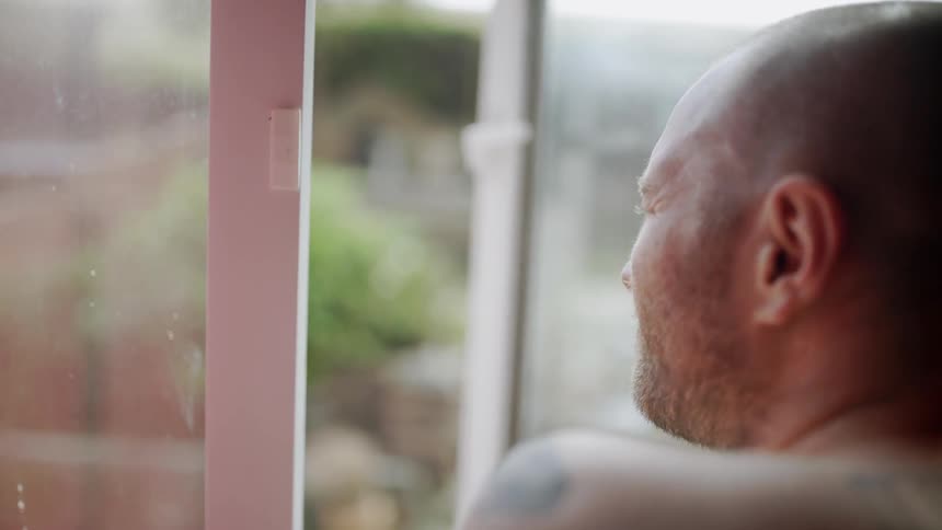 Shirtless man looks out the window