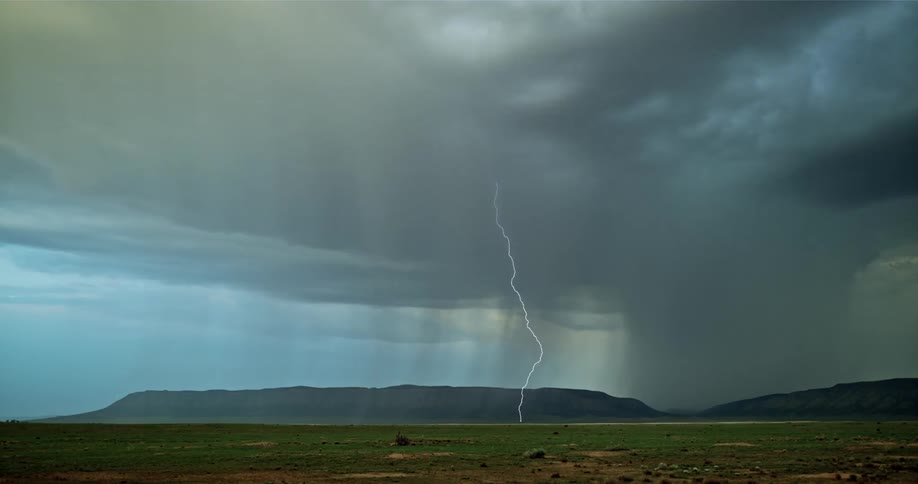 Lightning is flashing amid storm clouds as they move across a landscape at dusk
