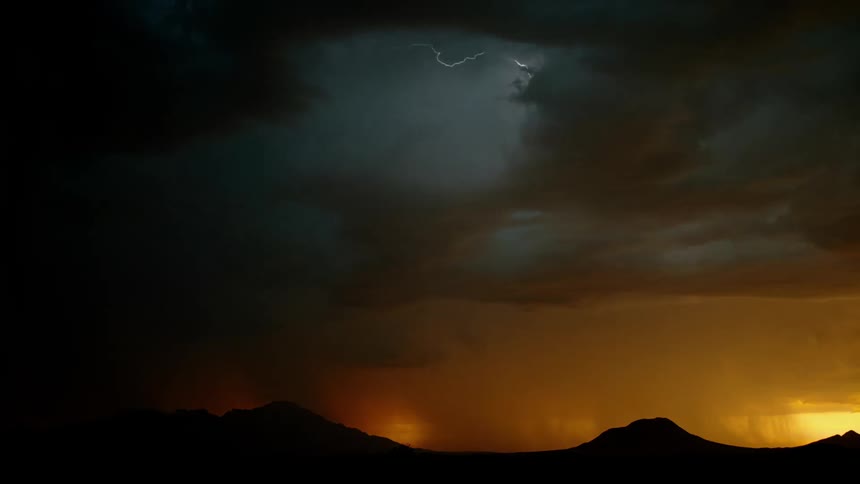 Lightning is flashing amid storm clouds as they move across a landscape at dusk