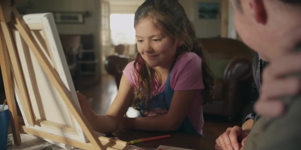A father talks to his daughter as she draws on a canvas sitting on an easel