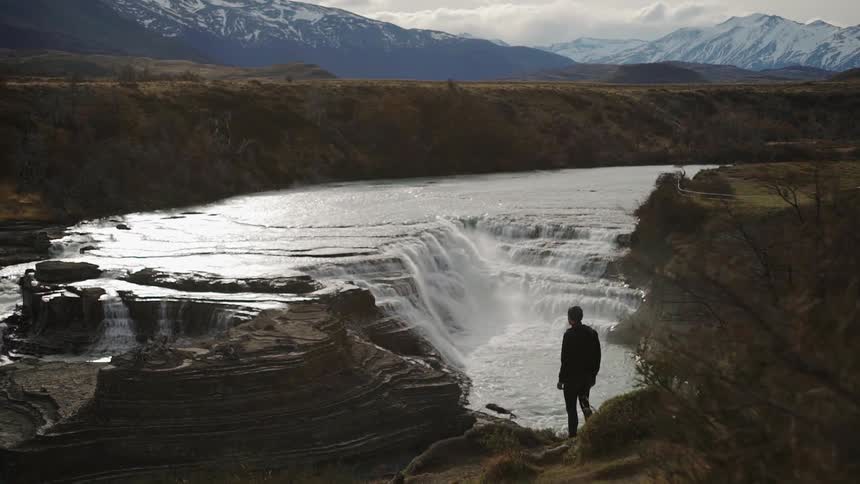 A man is standing on a ledge taking in a view of a waterfall in daylight