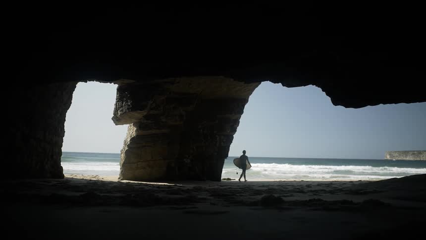 A man carries a surfboard past the rocks to the ocean