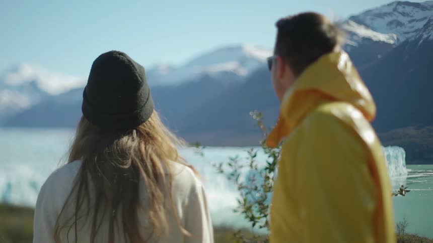 A man and woman are turning to look at a glacier outside on a sunny day