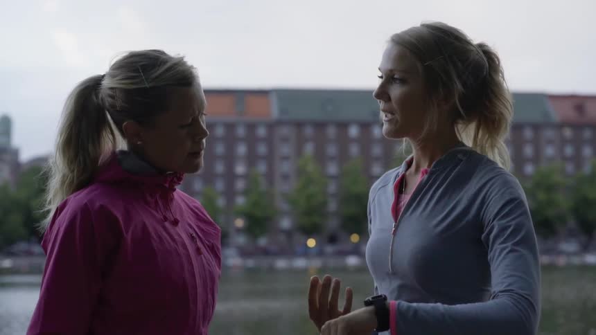 Two athletic friends converse together near water with city buildings in the background.