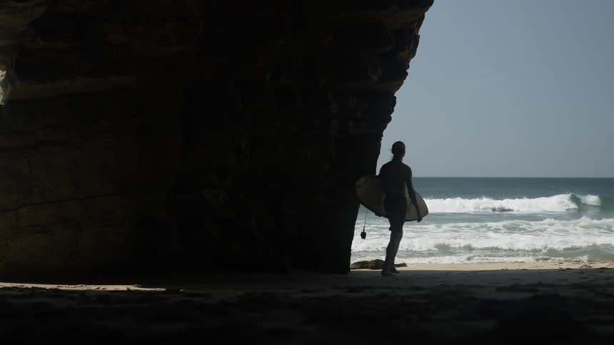 A surfer carries his board past rocks to the ocean