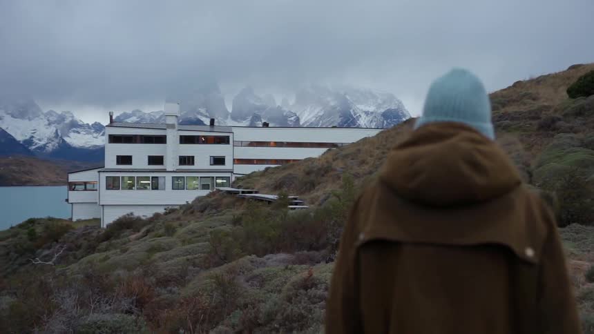 Person walks on hilly shore near water towards a building