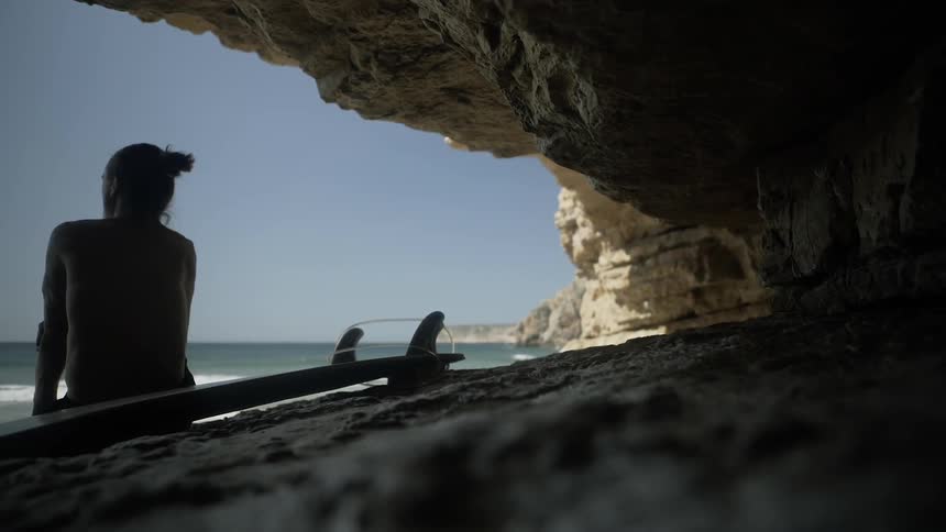 A surfer is sitting in the shade of a cave watching the ocean on a sunny day