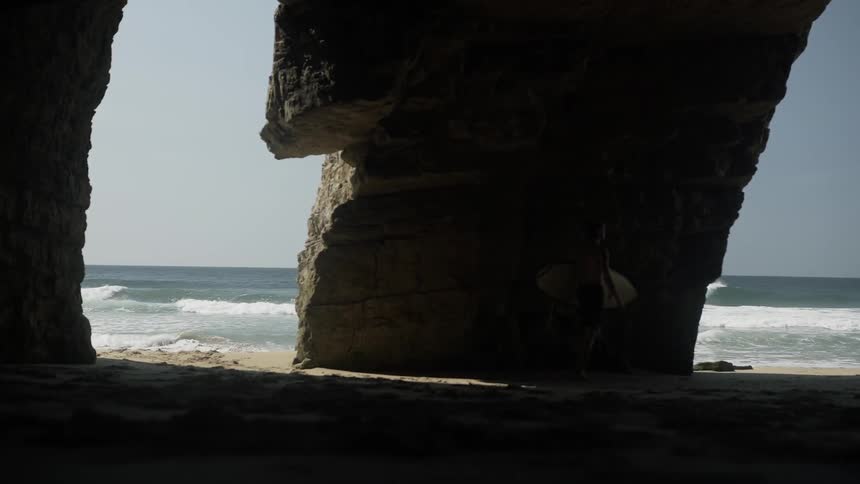 A surfer passes under a rock formation carrying his board