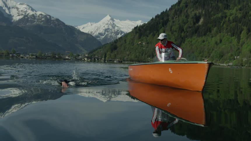 Person swims in a river alongside a man sitting in a small orange boat
