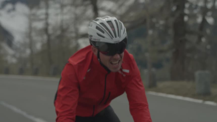 Man wearing a red coat pedals a bike on a country road in the rain