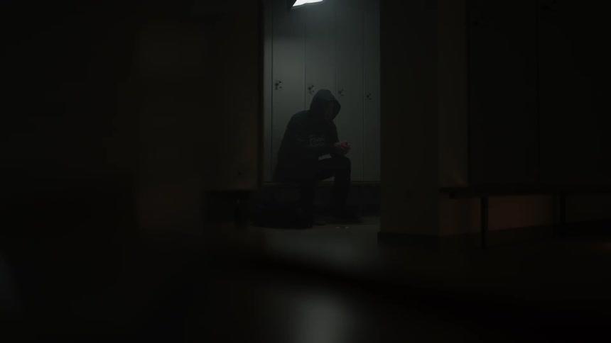 A man sits on a bench in front of lockers with his hands clasped