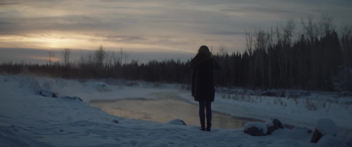 A woman is standing in the snow next to a small pond of water