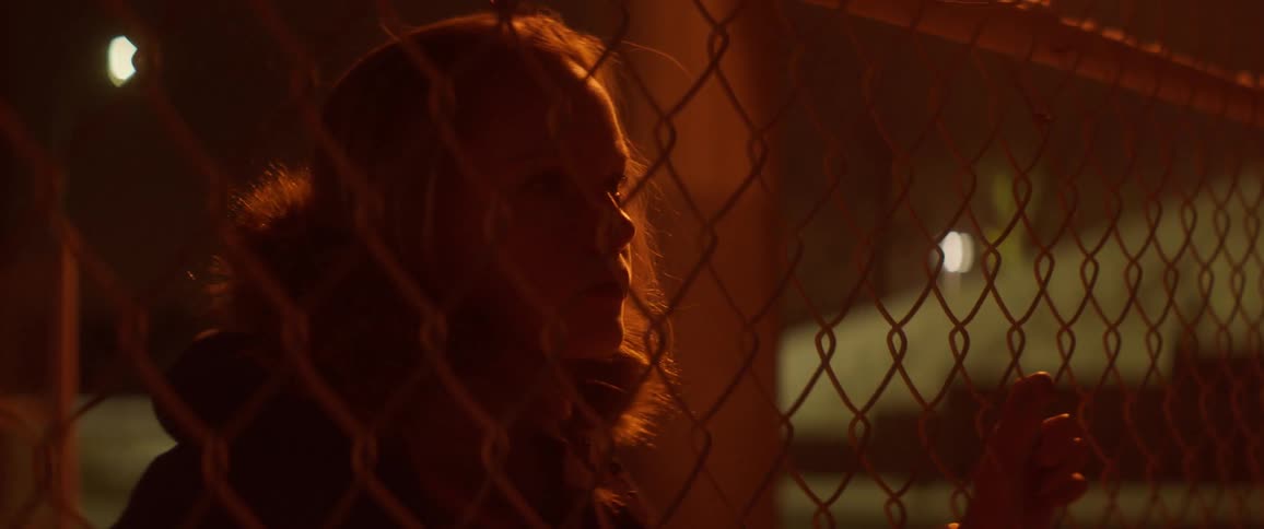 A teenage girl is removing her jacket hood as she peers through a fence at night