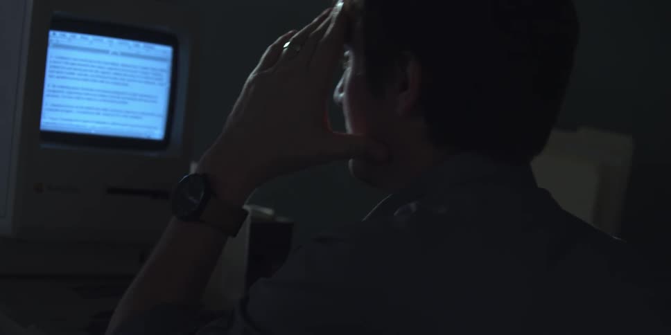 A man sitting at a desk reading information off a computer monitor before resting his head in his hand