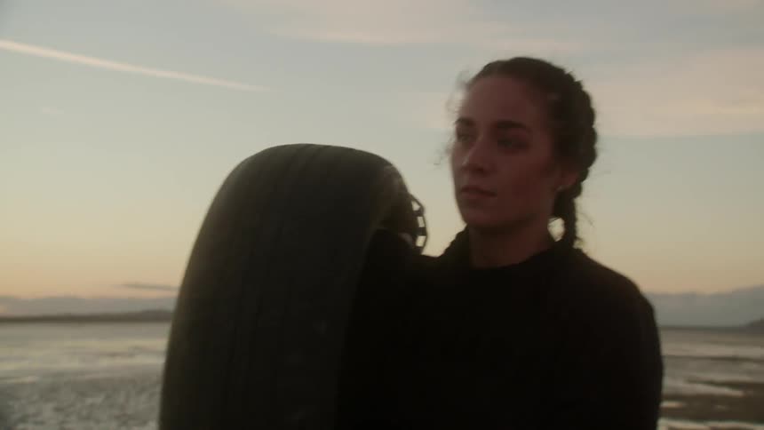 A woman is carrying a tire as she walks on a beach at dusk