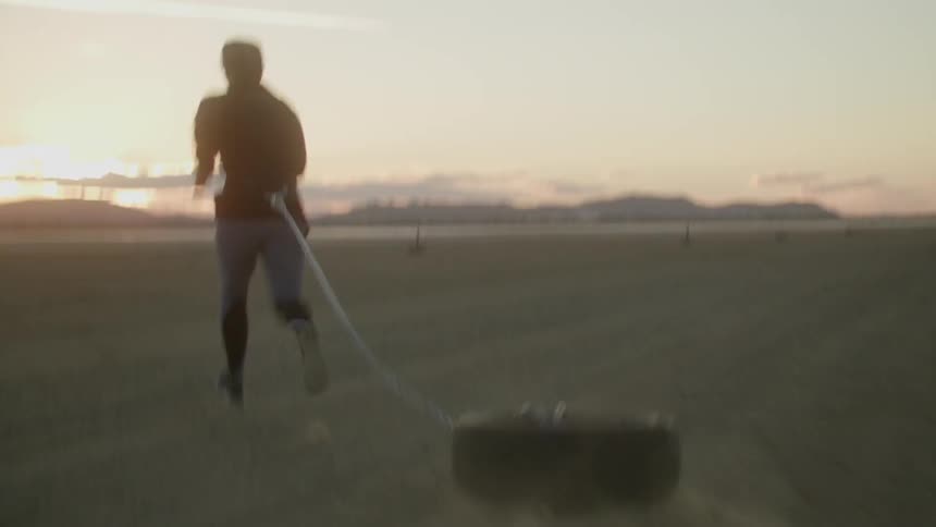 A woman is dragging a tire as she runs across a beach at sunset