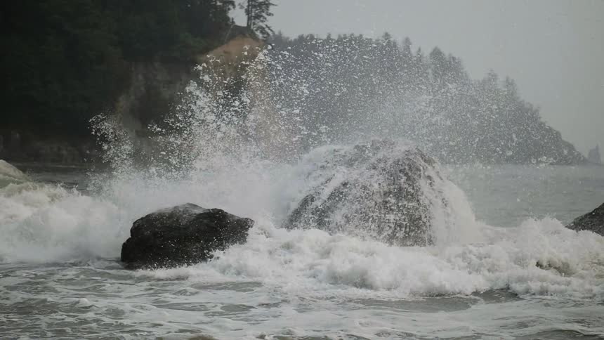 A wave is crashing against rocks as it flows toward a beach in daylight