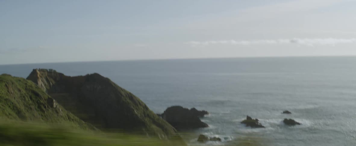 A vehicle is driving on a coastal road with a view of the ocean on a sunny day