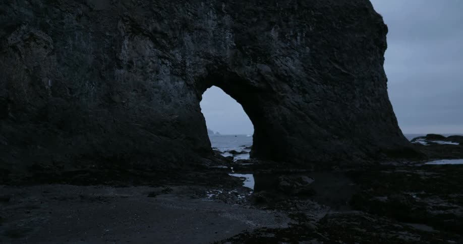 Ocean waves are flowing toward an arch in a cliff on a beach at dusk