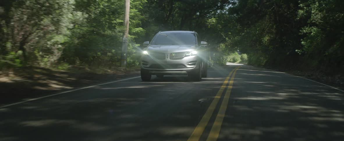 A vehicle is driving on a rural road with its lights on in daylight