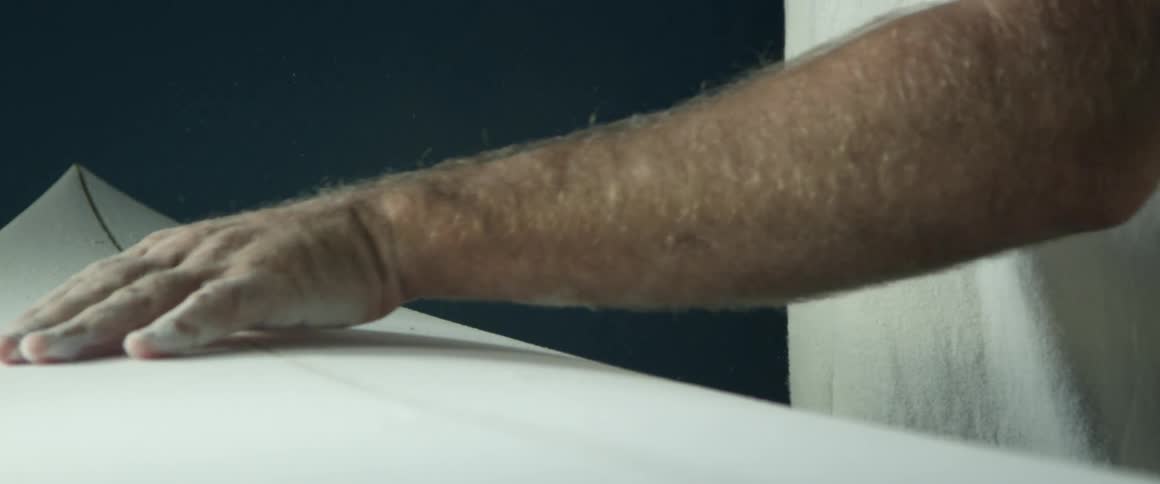 A white man rubs shavings off of a surfboard