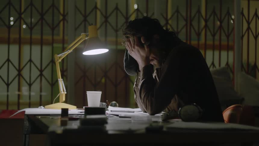 A man is rubbing his head as he works at a desk at night