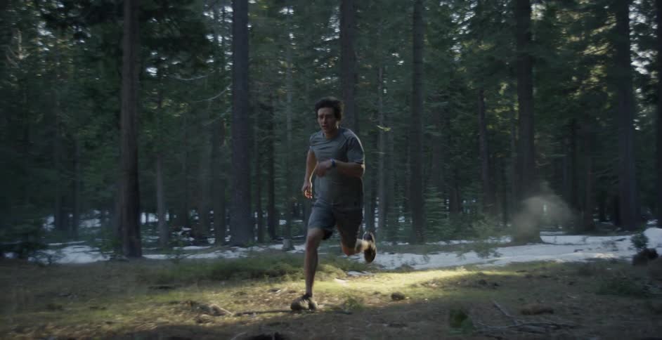 A man in shorts is running across a snow-covered forest floor in daylight