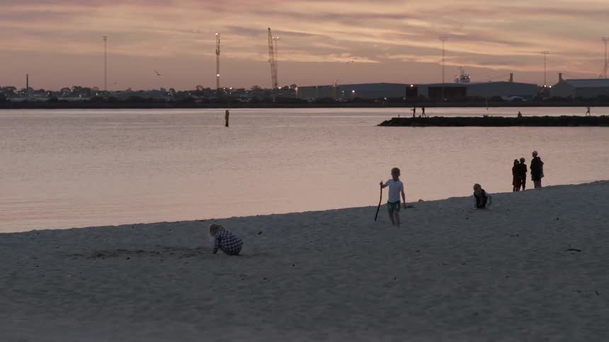 Several young children play on the beach while their parents stand together some distance away