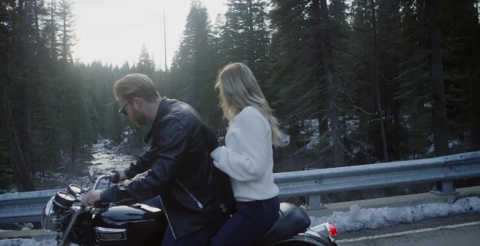 A couple riding a motorcycle are stopped looking at a river in a forest in winter